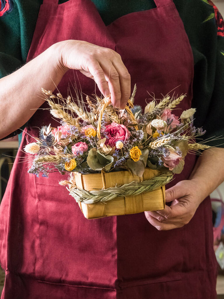 Kukkakauppias käsissä kukkakimpun kuivattuja kukkia ruusu, vehnä, eukalyptus paju kori kahvat, koristeltu viikate ruohoa. Taustaa esiliina kukkakauppias fuksia väri - Valokuva, kuva