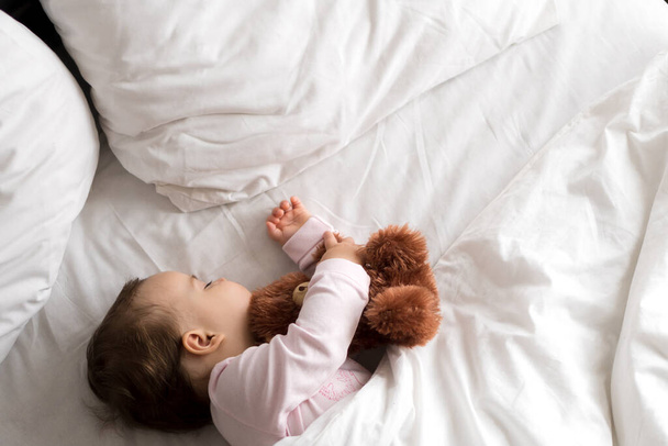 Αυθεντικό πορτρέτο χαριτωμένο καυκάσιο μικρό παχουλό κοριτσάκι ή αγόρι σε ροζ ύπνο με αρκουδάκι στο λευκό κρεβάτι. το παιδί ξεκουράζεται το μεσημέρι. φροντίδα, Υπνηλία, Παιδική ηλικία, Parenthood, έννοια της ζωής - Φωτογραφία, εικόνα