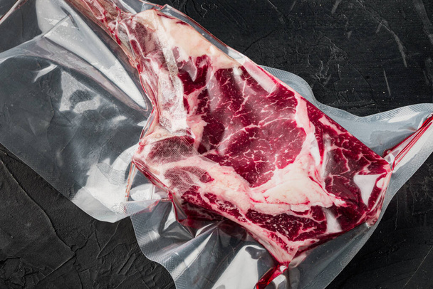 Dry aged raw tomahawk beef steak in plastic airtight pack set, on black stone background - Foto, Imagem