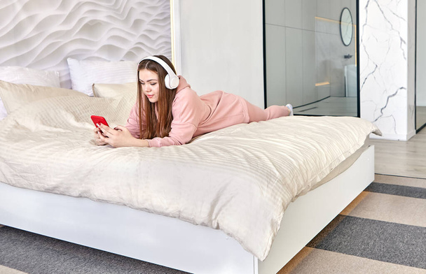 Young woman 20 years old lies in bed on her stomach and listens to music through headphones. - Foto, Bild