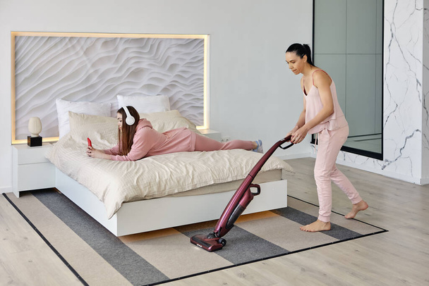 Using cordless vacuum cleaner to clean bedroom, mother does cleaning while daughter listens to music. - Foto, Imagen