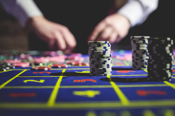 A close-up vibrant image of multicolored casino table with roulette in motion, with casino chips. the hand of croupier, money and group of gambling rich wealthy people playing in the background  - 写真・画像