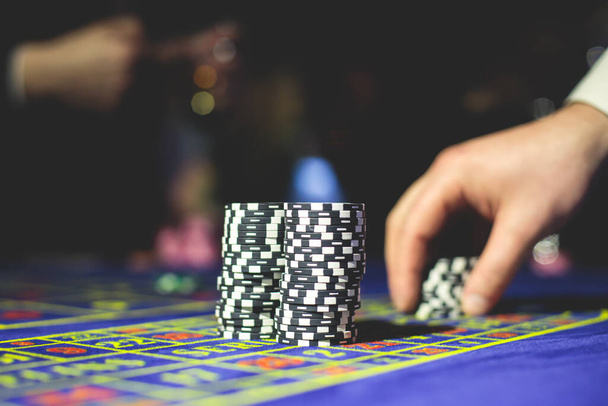 A close-up vibrant image of multicolored casino table with roulette in motion, with casino chips. the hand of croupier, money and group of gambling rich wealthy people playing in the background  - Photo, image