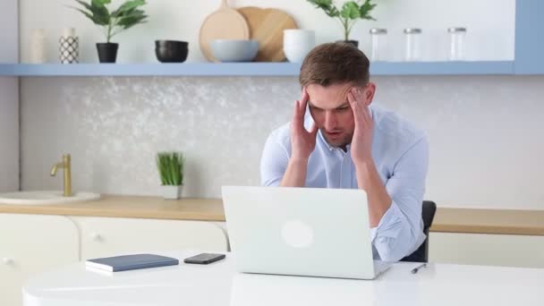 Bir adam elinde dizüstü bilgisayarla masada oturuyor ve kafasını tutuyor. Düzensiz çalışma günü. İş yorgunluğu. İş adamı kötü haberi okudu. İş sorunları. - Video, Çekim