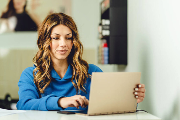 Heureuse femme brune caucasienne secrétaire en utilisant un ordinateur portable au travail à son bureau en ajustant l'écran en prenant rendez-vous de vraies personnes concept de petite entreprise et d'entrepreneur - Photo, image