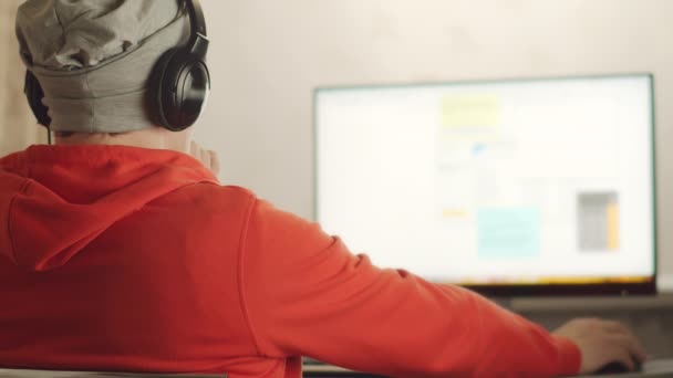 Un uomo con cappello grigio e cuffie lavora su un computer, riflessione sul muro, lavoro a distanza - Filmati, video