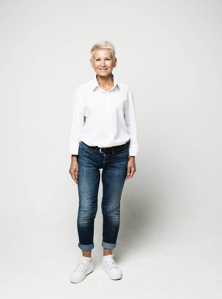 Full length photo of stylish elderly woman wearing jeans and white blouse with short white hair posing on a light gray background. - Фото, зображення
