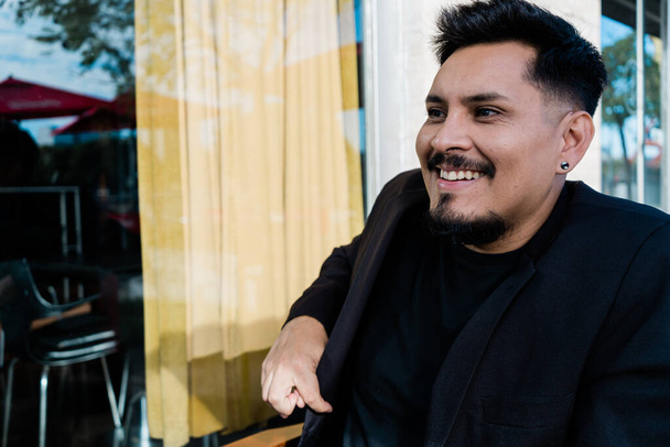 Egy fiatal latin férfi ül egy bárban és mosolyog. Fekete öltöny. - Fotó, kép