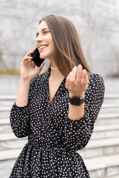 Vertikální portrét radostné podnikatelky, která mluví do mobilního telefonu s úsměvem venku, artikuluje paží - Fotografie, Obrázek