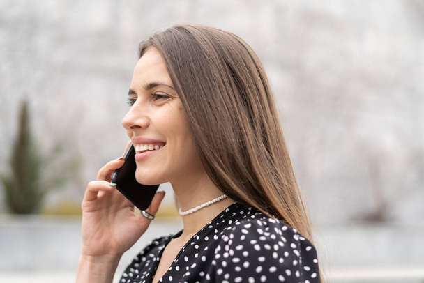 Πορτρέτο μιας εκθαμβωτικής καυκάσιας γυναίκας που μιλάει στο κινητό τηλέφωνο με καστανά μαλλιά φορώντας φόρεμα. Κοιτάζει αλλού με ένα τέλειο χαμόγελο, αντιγράφει το χώρο. - Φωτογραφία, εικόνα