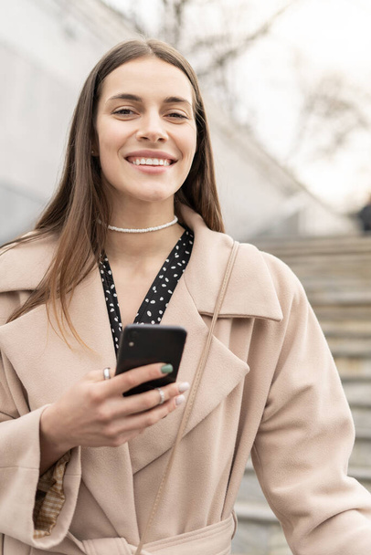 Portrait of a fashionable young woman holding mobile phone wearing coat and smiling in to the camera - Foto, Imagen