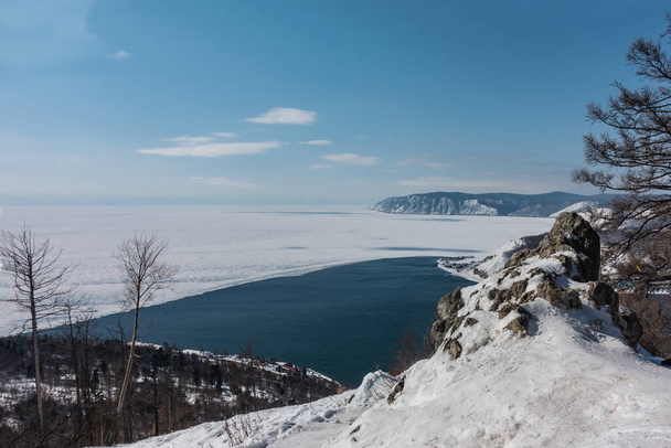 Rzeka Angara wypływa z jeziora Baikal. Granica między lodem a niebieską, niezamarzającą wodą. Na pierwszym planie znajduje się malowniczy kamień pokryty śniegiem. W oddali jest pasmo górskie.. - Zdjęcie, obraz