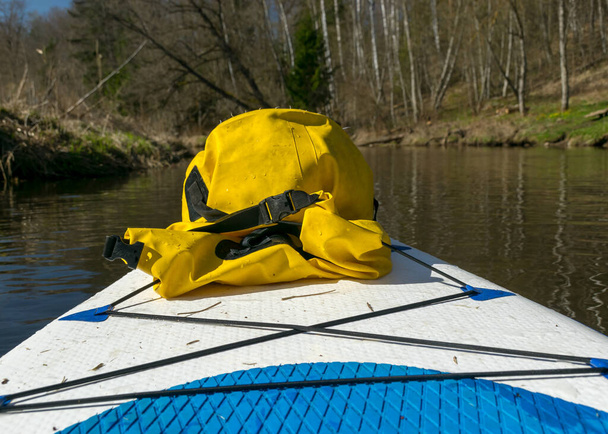 Landschaft mit einem gelben wasserdichten Sack im Vordergrund auf einem Paddelbrett, im Hintergrund ein Blick auf die Ufer eines kleinen Wildbaches im Frühling - Foto, Bild