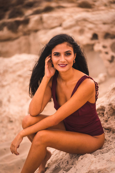 Portrait of a young brunette Caucasian woman in a maroon swimsuit, sitting smiling on the beach of Cabo de Gata, Nijar. Andalucia, Spain - Photo, Image