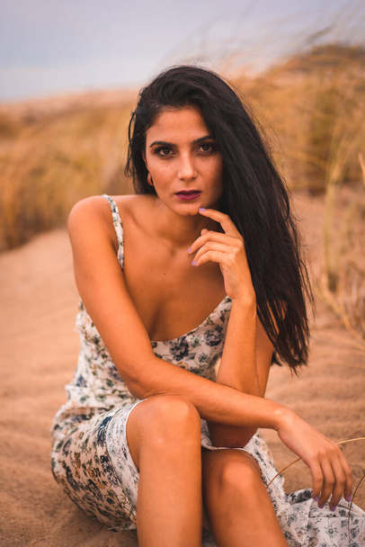 Portrait of a brunette Caucasian caressing her hair in a white floral dress, sitting on the sand on the beach of Cabo de Gata, Nijar. Andalucia, Spain - Foto, imagen