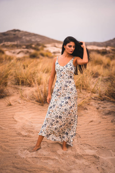 Portrait of a Caucasian brunette stroking her hair in a white floral dress, in the sand on the beach of Cabo de Gata, Nijar. Andalucia, Spain - Φωτογραφία, εικόνα