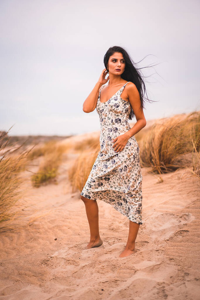 Portrait of a Caucasian brunette stroking her hair in a white floral dress, in the sand on the beach, vertical photo - Fotografie, Obrázek