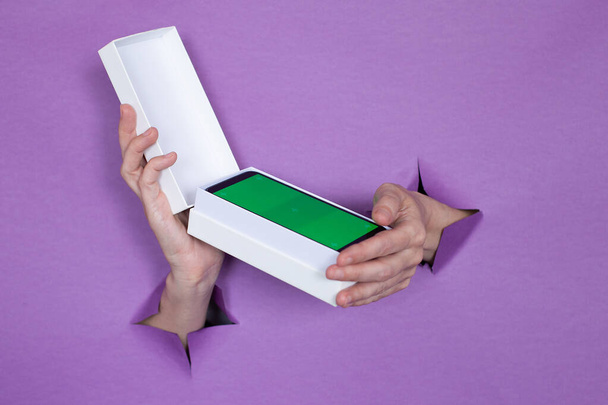 un nouveau smartphone avec un écran vert dans un paquet sur fond violet, une fille tient dans ses mains, des trous dans l'arrière-plan. - Photo, image