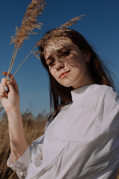 Улыбающаяся молодая женщина в белой блузке, стоящая в поле и держащая в руках ветки сухой травы пампасов перед небом. Стиль и мода. Девушка в повседневной одежде с крупным планом. Золотой час - Фото, изображение