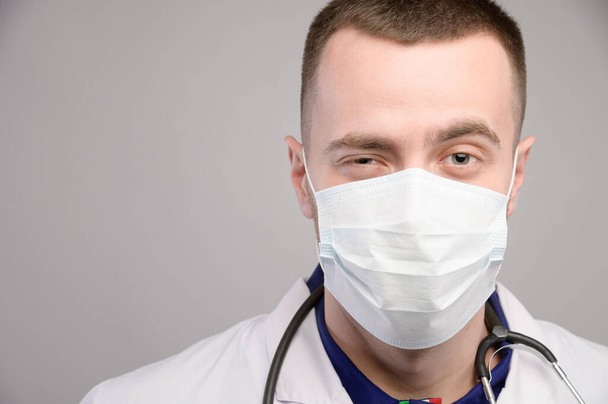 Portrait of a young friendly friendly Caucasian doctor in a protective medical mask on a gray background. A man winks and smiles with his eyes looking at the camera - Photo, image