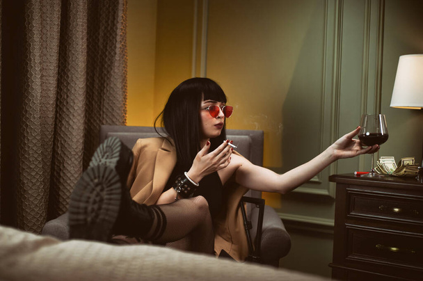 Egy női bűnöző egy hotelben ül egy széken, cigarettázik és bort iszik. - Fotó, kép