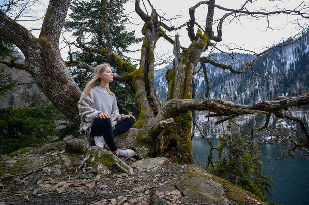 Dziewczyna siedzi na drzewie i medytuje na tle błękitnego jeziora. Jezioro Ritsa w Abchazji - Zdjęcie, obraz