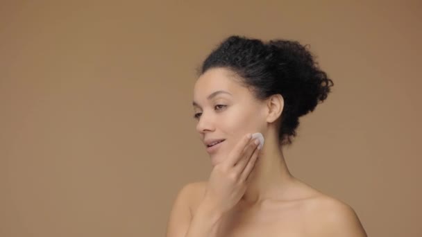 Beauty portrait of young African American woman cleansing face with white cotton pad, removing makeup. Black female on brown studio background. Side view. Slow motion ready, 4K at 59.94fps. - Video