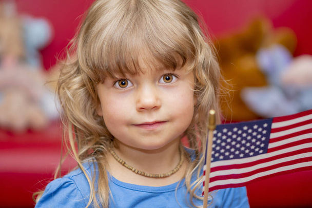 Porträt eines kleinen lächelnden blonden Mädchens mit amerikanischer Flagge auf neutralem Hintergrund. - Foto, Bild