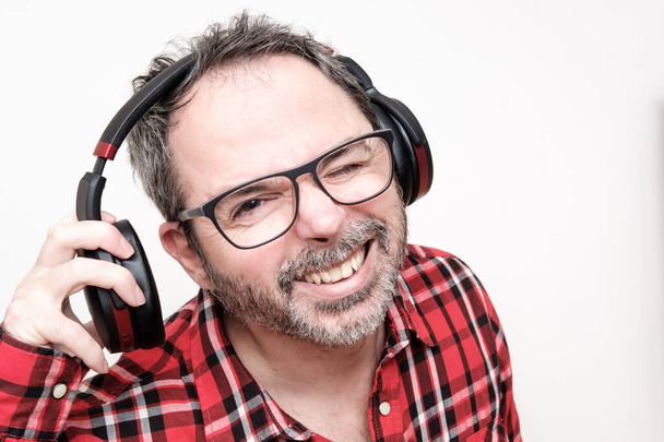 Portrait of a laughing mature man with beard wearing red plaid shirt and glasses. Using headphones. - Photo, Image