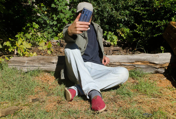 若い大人が草の上に座っている彼の携帯電話を見てカジュアルな服と赤いエスパドリルとグレーの帽子と緑のジャケット - 写真・画像