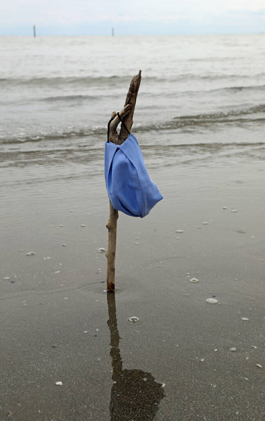 хирургическая маска на берегу моря, висящая на палочке, как флаг на полу-мачте - Фото, изображение