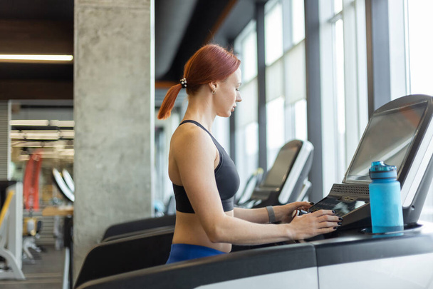 Portrait d'une femme aux cheveux roux dans une salle de gym moderne. Fit femme mince travaille sur un tapis roulant, entraînement cardio, mode de vie sain. - Photo, image