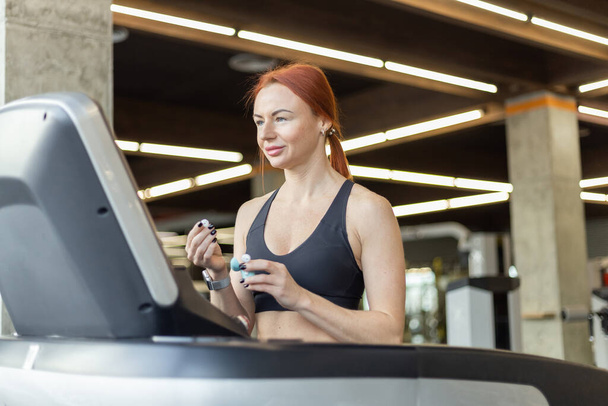 Portrait of a red-haired fitness woman with earphones in a modern gym. Fit slim woman works out on a treadmill, cardio workout, healthy lifestyle. - Фото, зображення