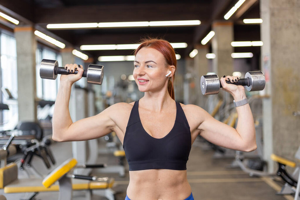 Attractive red-haired fitness woman trains shoulder muscles doing overhead dumbbell press in a modern gym. Bodybuilding and Fitness - Fotoğraf, Görsel