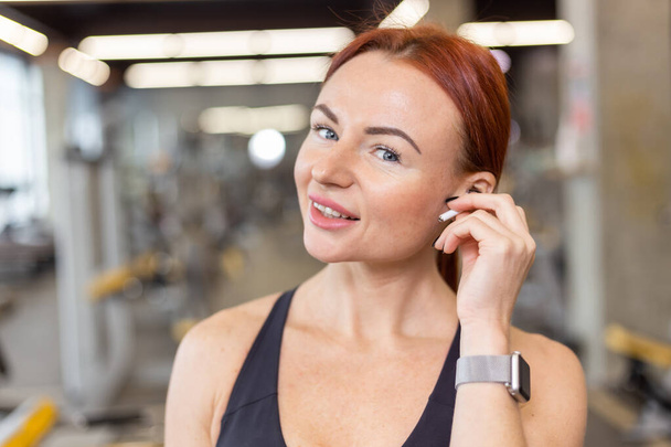Portrait of red-haired fitness woman with wireless headphones listening to music in a modern gym - Fotoğraf, Görsel