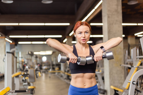 Привлекательная рыжеволосая фитнес-женщина тренирует мышцы плеч гантелями в современном тренажерном зале. Культуризм и фитнес - Фото, изображение