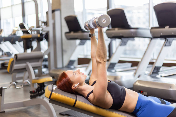 Die athletische rothaarige Frau trainiert die Brustmuskulatur mit Hanteln in den Händen, während sie auf einer Schrägbank liegt. Workout im Fitnessstudio - Foto, Bild