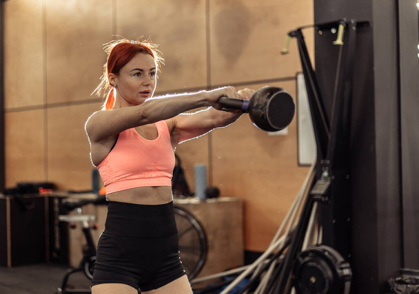 Modern bir spor salonunda elinde kettlebell olan kızıl saçlı bir kadının yoğun egzersizi. - Fotoğraf, Görsel