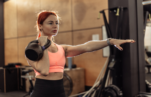 Intensives Training einer rothaarigen Frau mit einer Kettlebell in der Hand in einem modernen Fitnessstudio - Foto, Bild