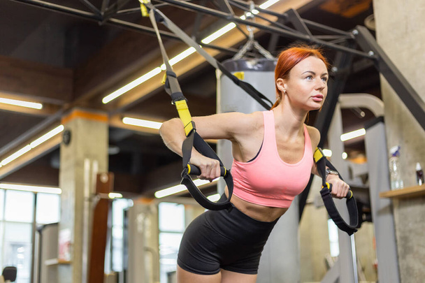 TRX Training. Junge athletische rothaarige Frau im Sportbekleidungstraining mit Trageschlaufen im Fitnessstudio. - Foto, Bild