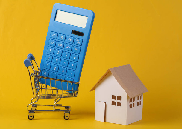Huisaankoop. Mini papieren huis model, rekenmachine in supermarkt trolley op gele achtergrond.  - Foto, afbeelding