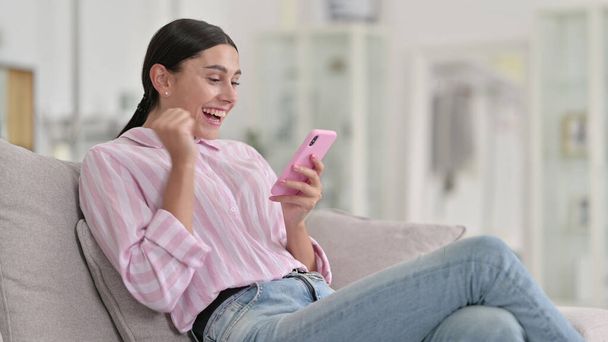 Excited Latin Woman Celebrating Success on Smartphone at Home  - 写真・画像