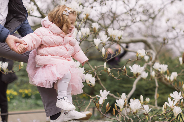 Happy young father holding his daughter's hand. Warmly dressed. Background of magnolia blossoms. - Foto, Imagem