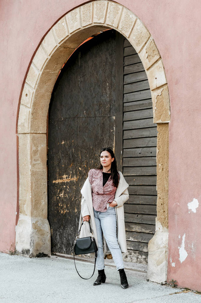 A stylish young brunette woman from Croatia standing in front of an old arch wooden door of a red building - Valokuva, kuva