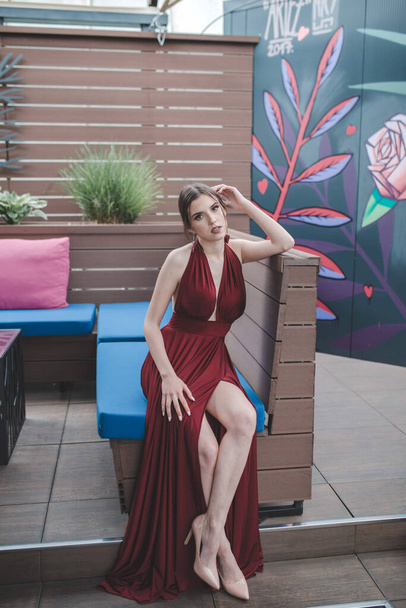 A vertical shot of a Caucasian attractive woman with a red sexy evening dress on the outdoor cafe - Фото, изображение
