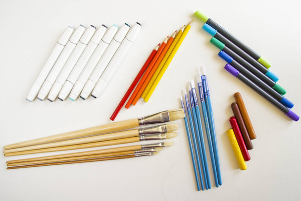 A top view of paintbrushes and crayons with colorful markers and pencils on a white surface - Fotó, kép