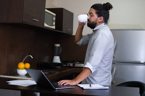 adulto joven tomando una bebida de pie relajado en la cocina mientras mira el portátil al lado de un portátil, trabajando en casa. - Foto, Imagen
