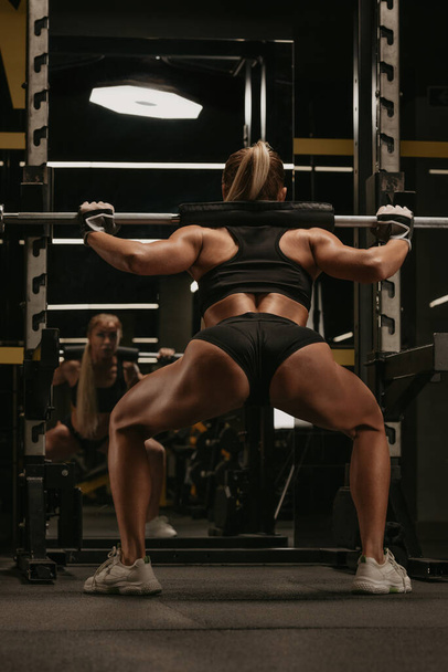Фото со спины спортивной женщины с светлыми волосами, которая сидит на корточках рядом со штангой в спортзале. Девушка занимается спортом на ногах. - Фото, изображение