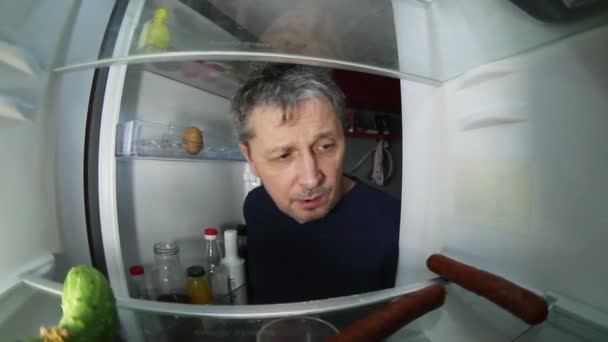 Un hombre sin afeitar con resaca busca en el refrigerador una bebida y algo de comer.. - Metraje, vídeo