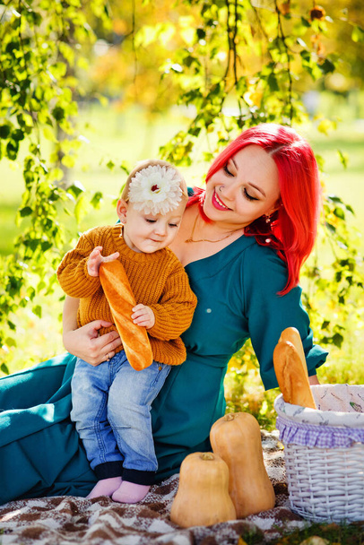 Matka z rudymi włosami przytula córkę, siedząc na kocu w parku na pikniku z bułką o zachodzie słońca. Koncepcja wakacji letnich. Dzień Matki, Dzień Dziecka. Spędzać razem czas. Promienie słońca. - Zdjęcie, obraz